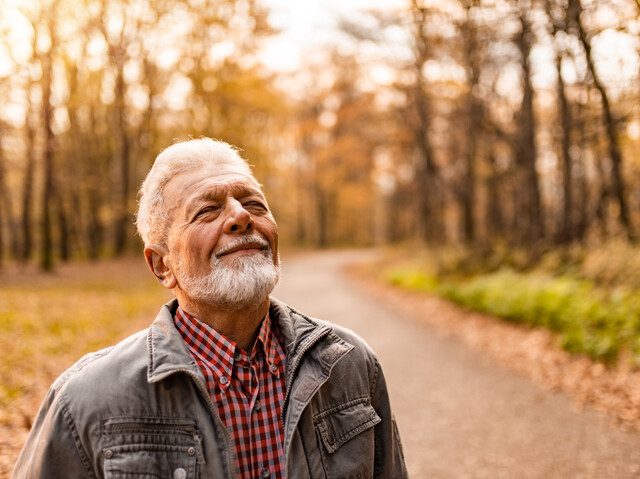 Tranquil senior man enjoying a walk on forest road on autumn day and breathing fresh air with eyes closed.