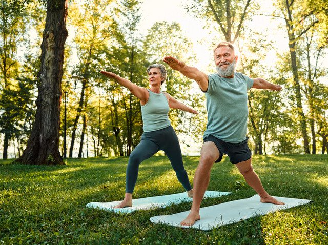 Determined family couple standing on rubber mats in national park and performing warrior asana pose. Aged man and woman showing strength and flexibility when practicing yoga.